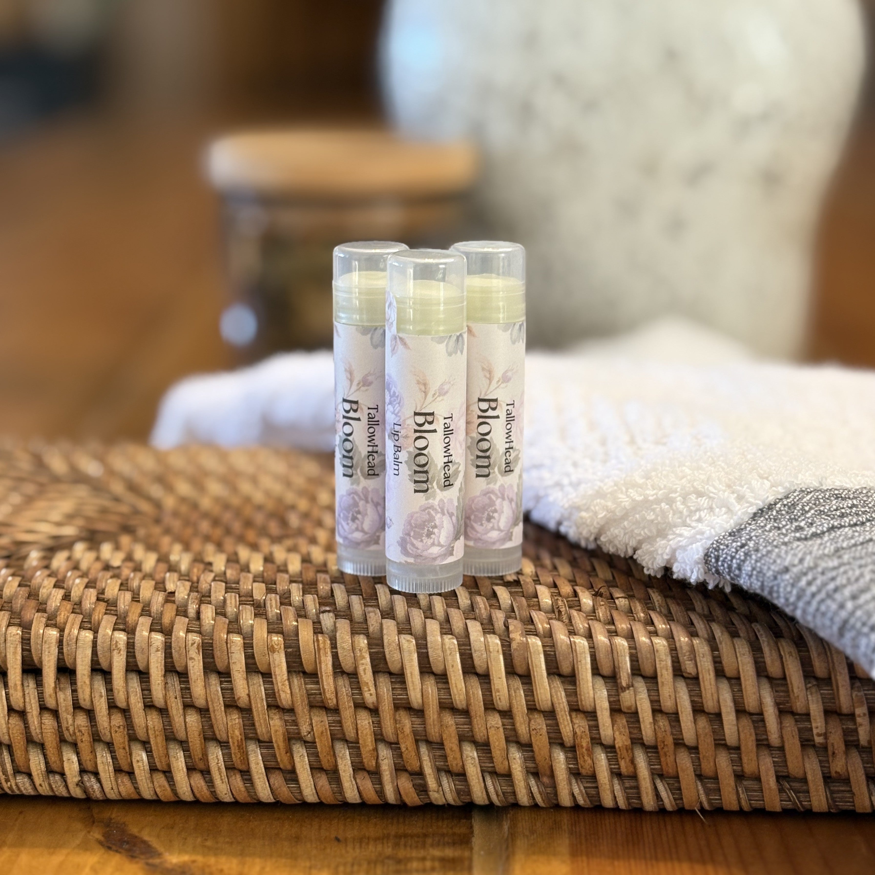Three lip balm tubes labeled 'Bloom' on a woven surface with a blurred background.