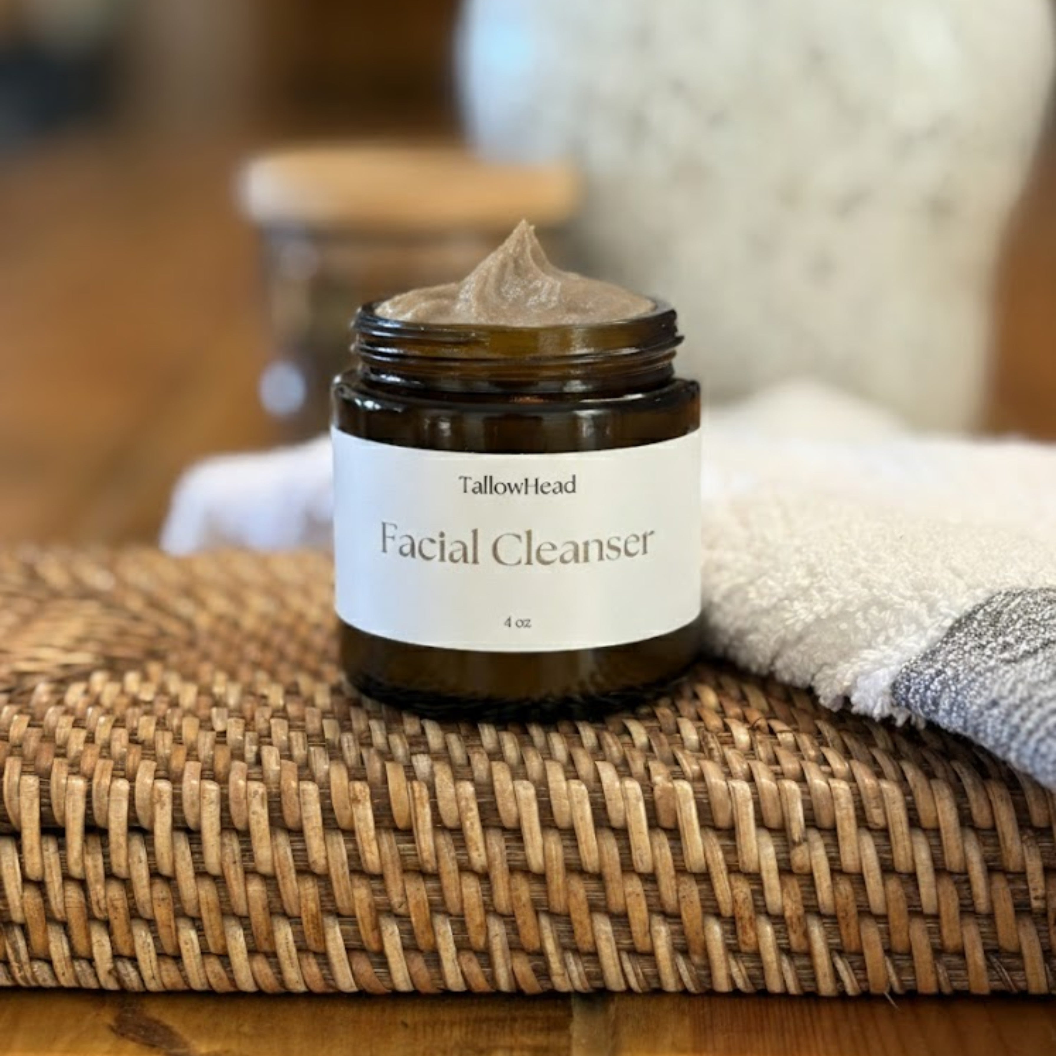 Jar of facial cleanser labeled 'TallowHead' on a woven mat with a towel and jar in the background.